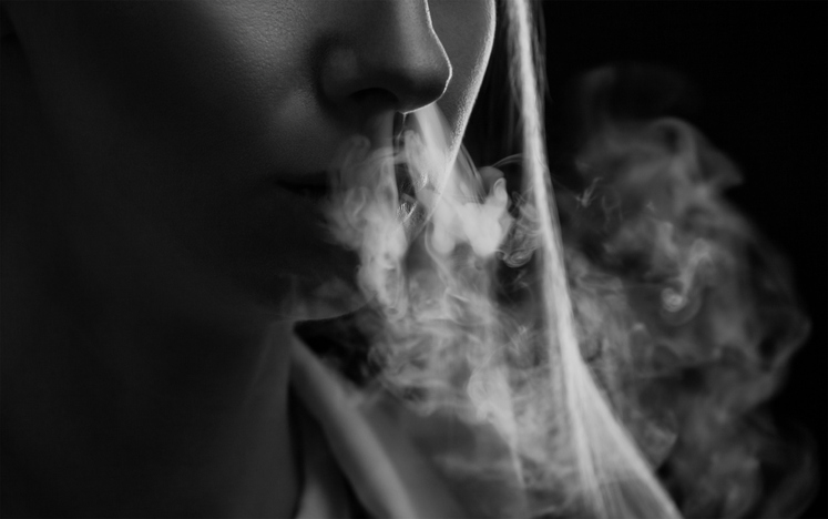 A young woman smokes cigarettes and blows smoke on a black background. Harm of smoking.