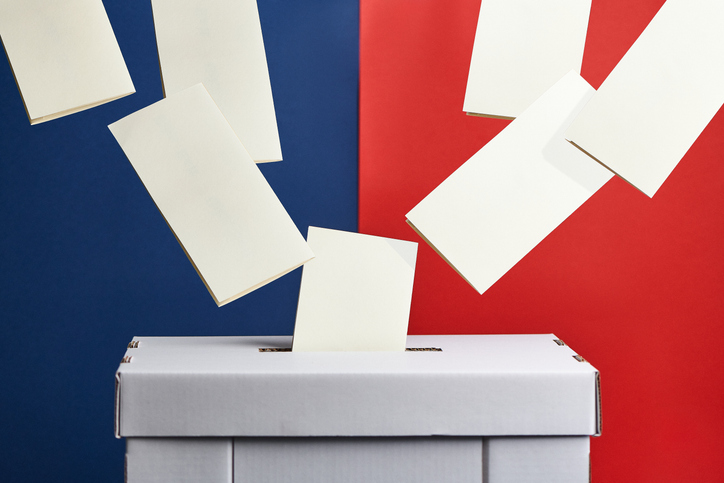 Cardboard ballot box with voter slips flying into it, with a blue and red coloured background.